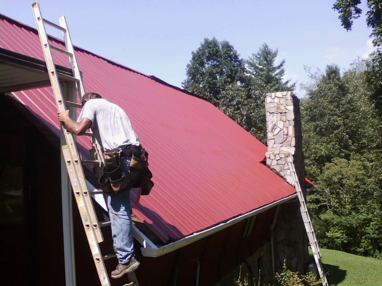 Roofer on a ladder beside a red metal roof and stone chimney — Soffit & Fascia Repair by Roofing Brothers in Etna Green